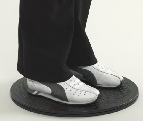 Pies de una persona con zapatillas blancas y pantalones negros, sobre una plataforma negra giratoria, sobre un fondo blanco.