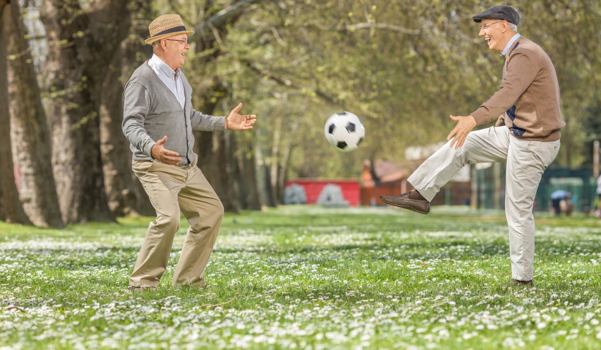 Dos hombres mayores juegan al fútbol en un parque con césped. Uno patea el balón, el otro se prepara para recibirlo.