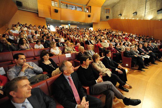 Público sentado en un teatro, presenciando un evento. Mucha gente, bien vestida, en una sala de tonos cálidos.