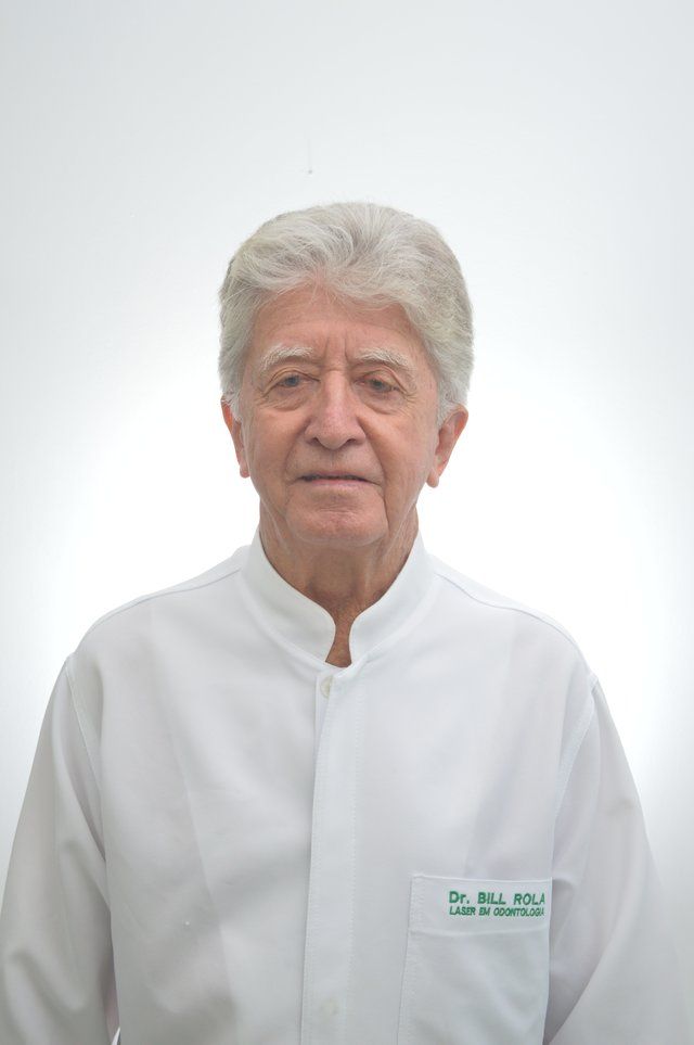 Pessoa idosa de jaleco branco, com cabelos grisalhos, olhando para a frente. No bolso está escrito “Dra. Bia Mello”.