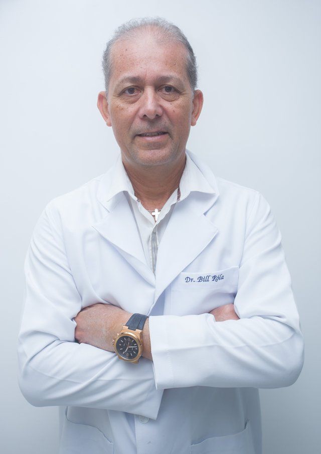Homem de jaleco branco, braços cruzados, relógio de ouro, em frente a um fundo branco.
