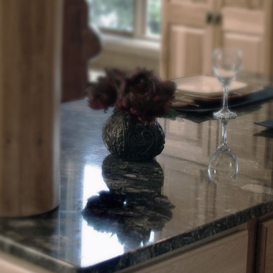 Small black vase with dark flowers on a glossy kitchen counter, reflected on the surface