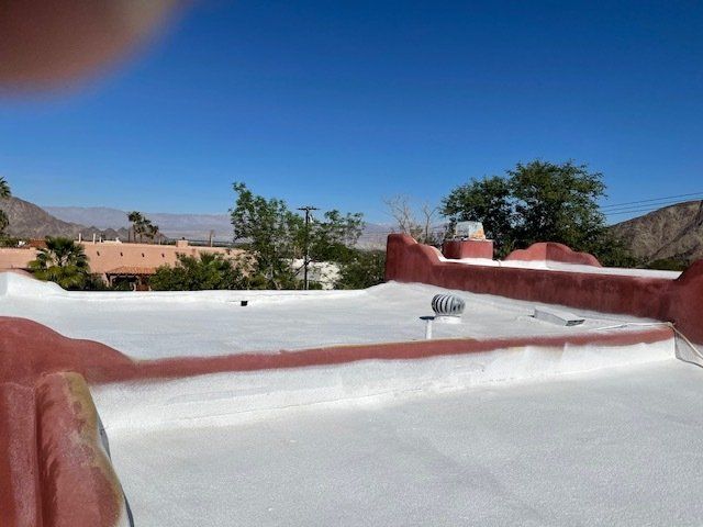 White-coated flat roof with red trim under a clear blue sky, overlooking houses and mountains.