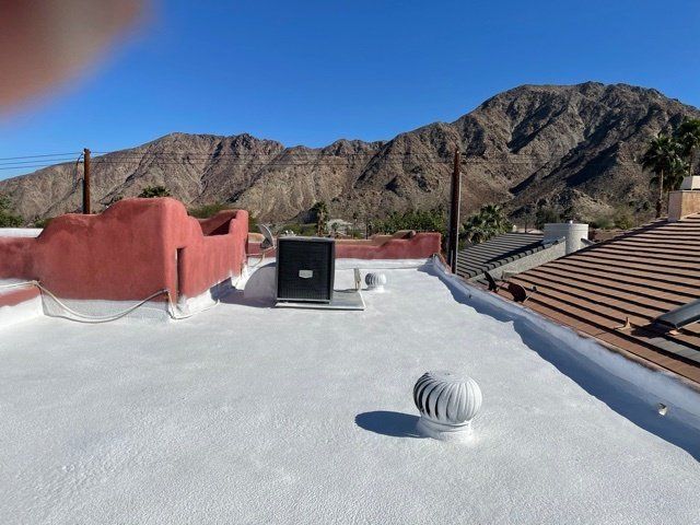 A flat roof with white coating, vents, and air conditioning unit against a mountain backdrop under a clear blue sky.