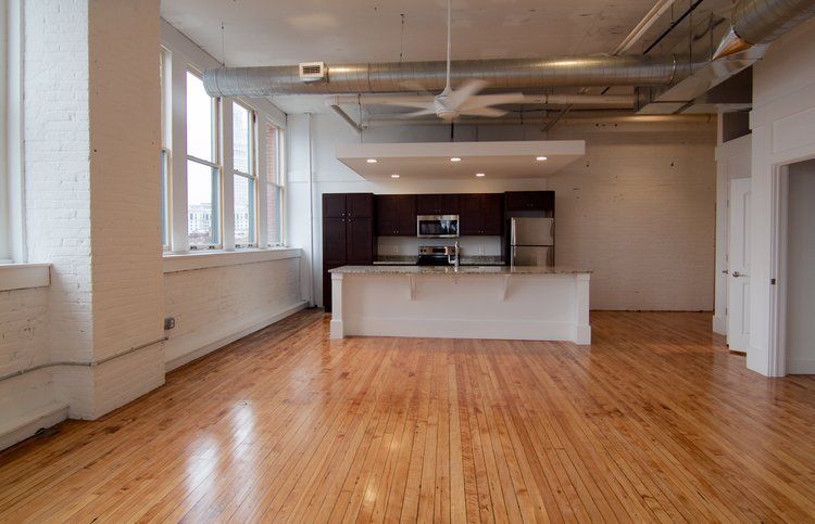 Empty loft-style room with hardwood floors, white walls, large windows, and a kitchen island at the back