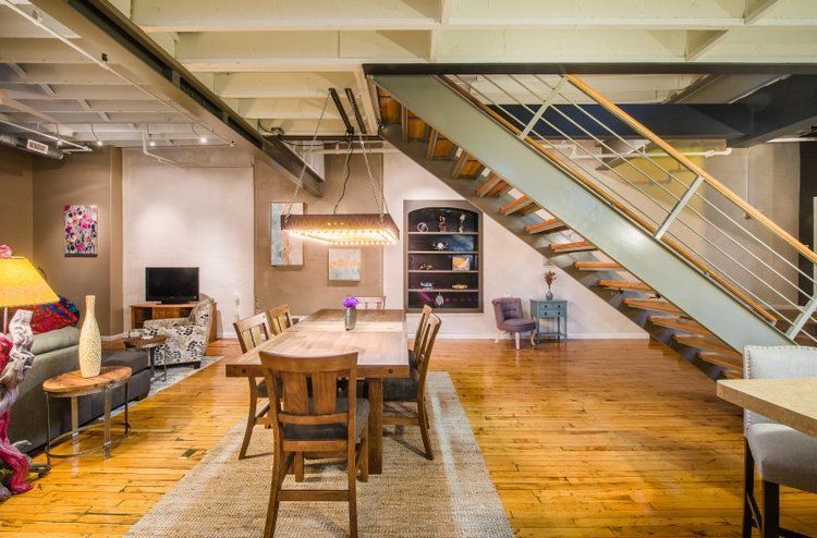Loft-style living room with wooden dining table, sofa seating, exposed beams, and staircase on the right