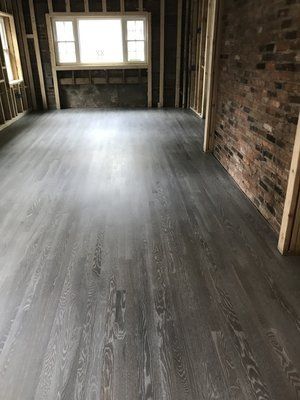 Empty unfinished room with dark wood flooring, exposed studs, brick wall, and a window at the far end.