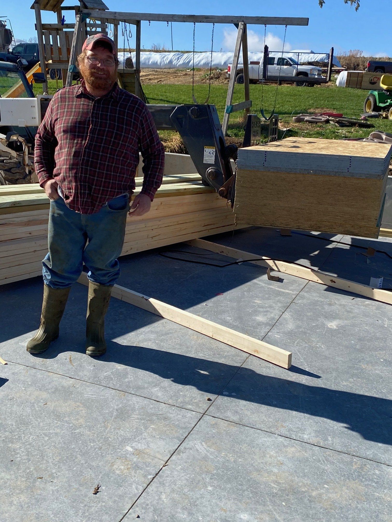 Man in boots and flannel stands near lumber on concrete, outdoors.