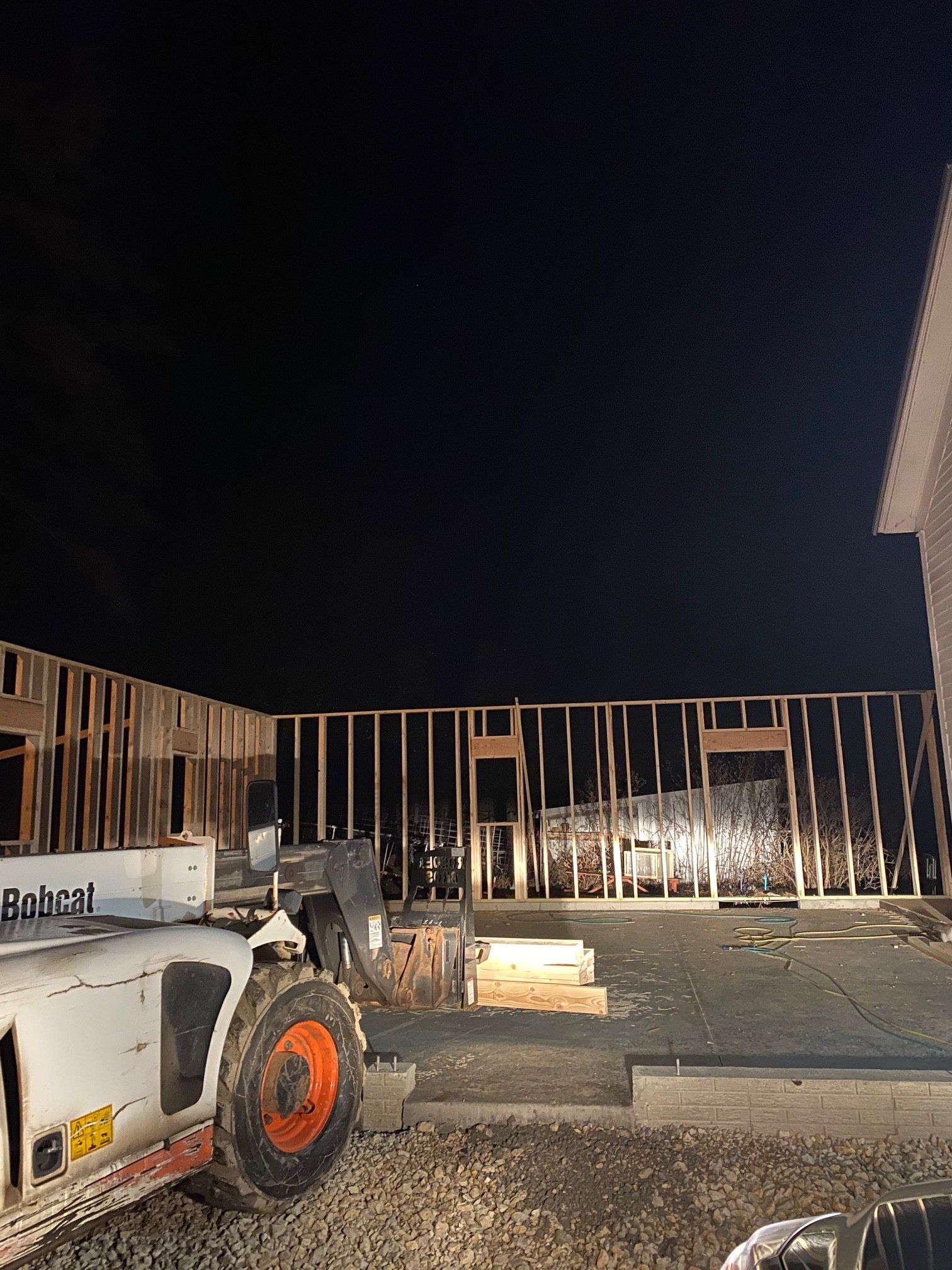 Night construction site: Bobcat loader in front of a partially framed building. Dark sky above.