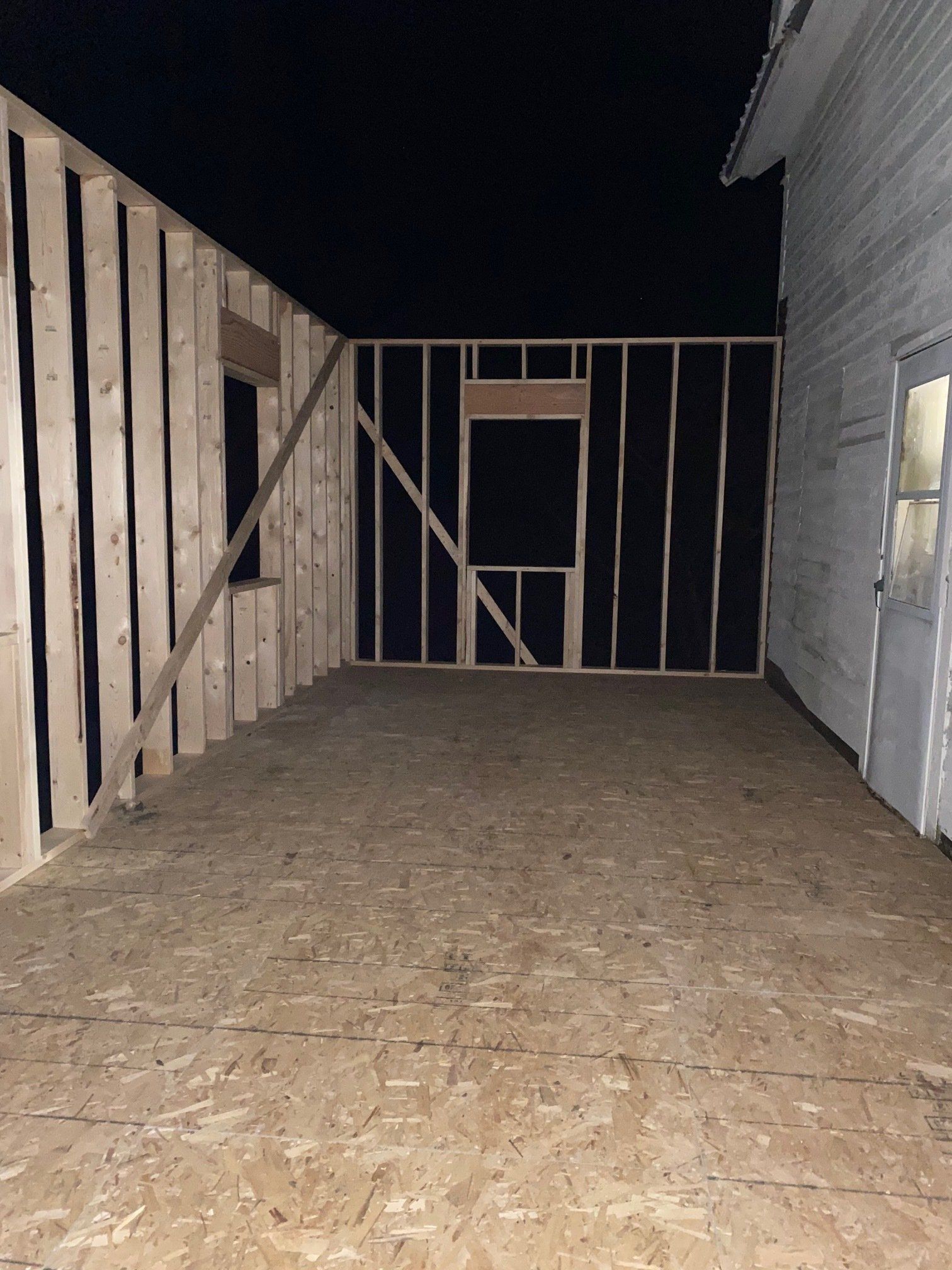 Framing of a room under construction with black insulation and a door to a white building.
