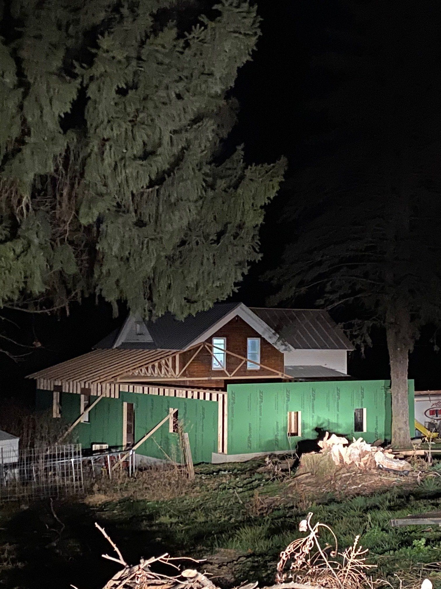 House under construction at night, green siding, partially lit. Tall trees frame the scene.