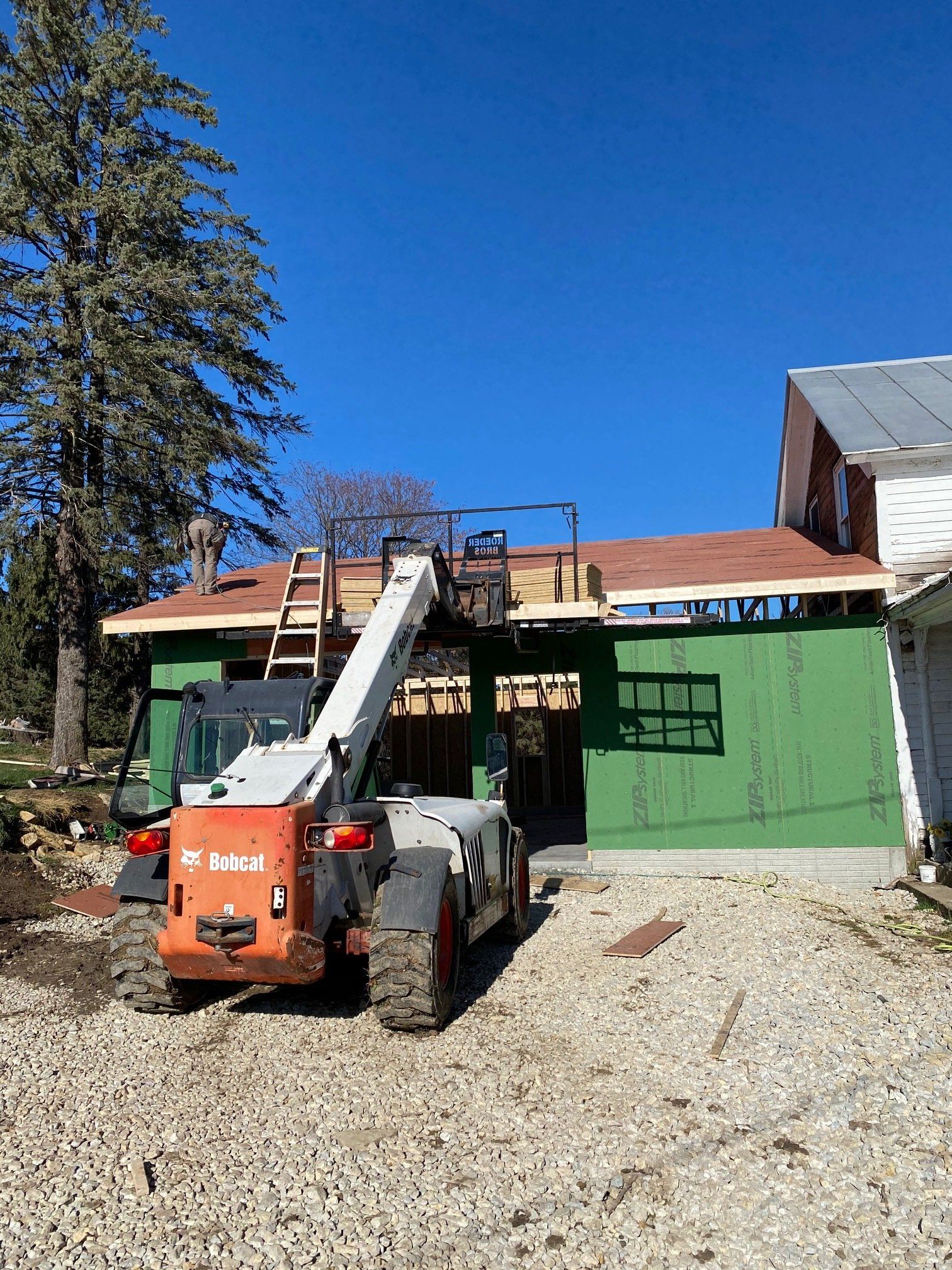 Bobcat telehandler at construction site; green siding, brown roof, blue sky.