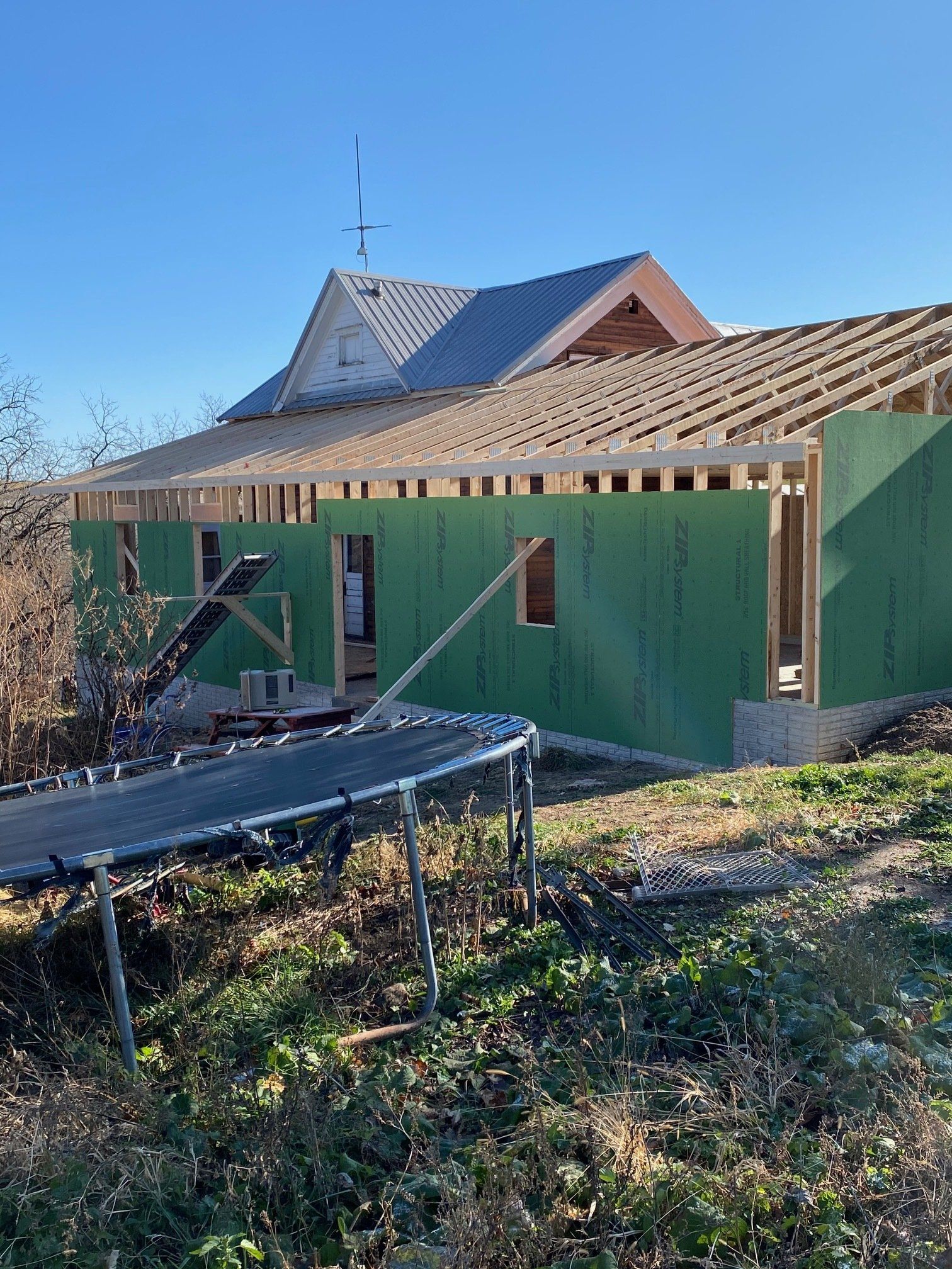 House under construction with green siding and partially built roof, trampoline in foreground.