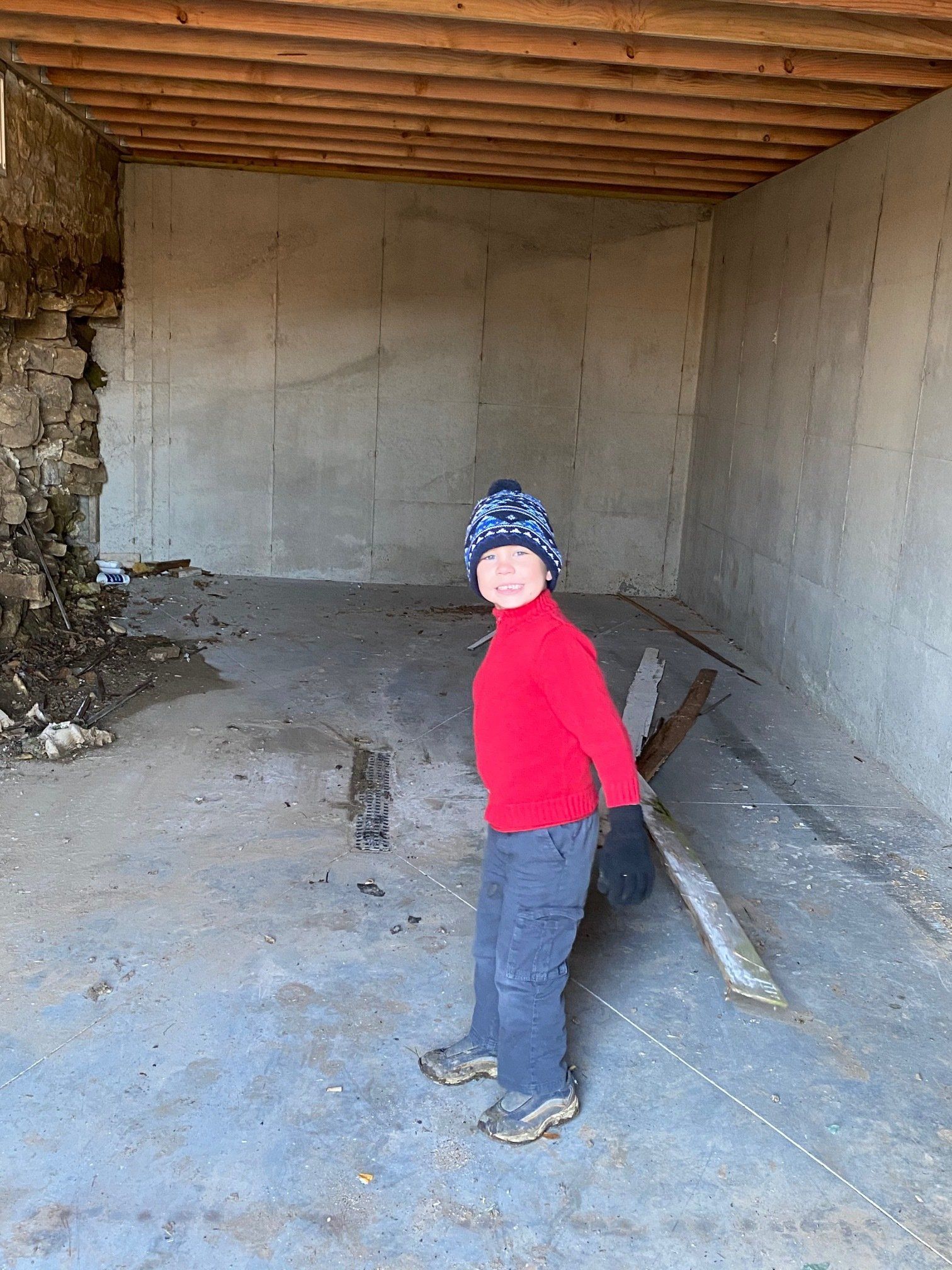 A child in a red shirt and blue hat smiles in a concrete room, holding a wooden plank.