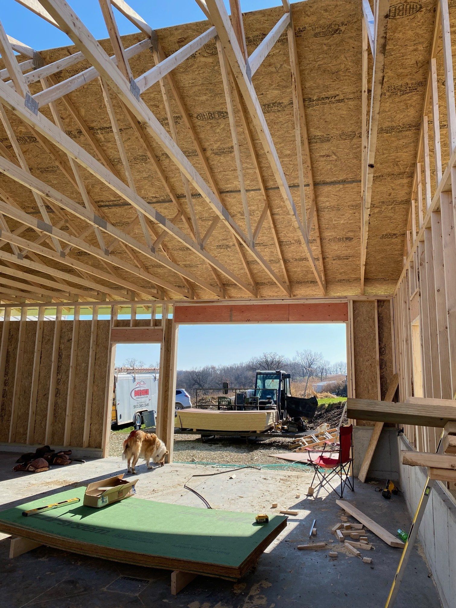 Construction site, wooden framing, open garage entrance, dog walks past, equipment and sunny outdoor view.