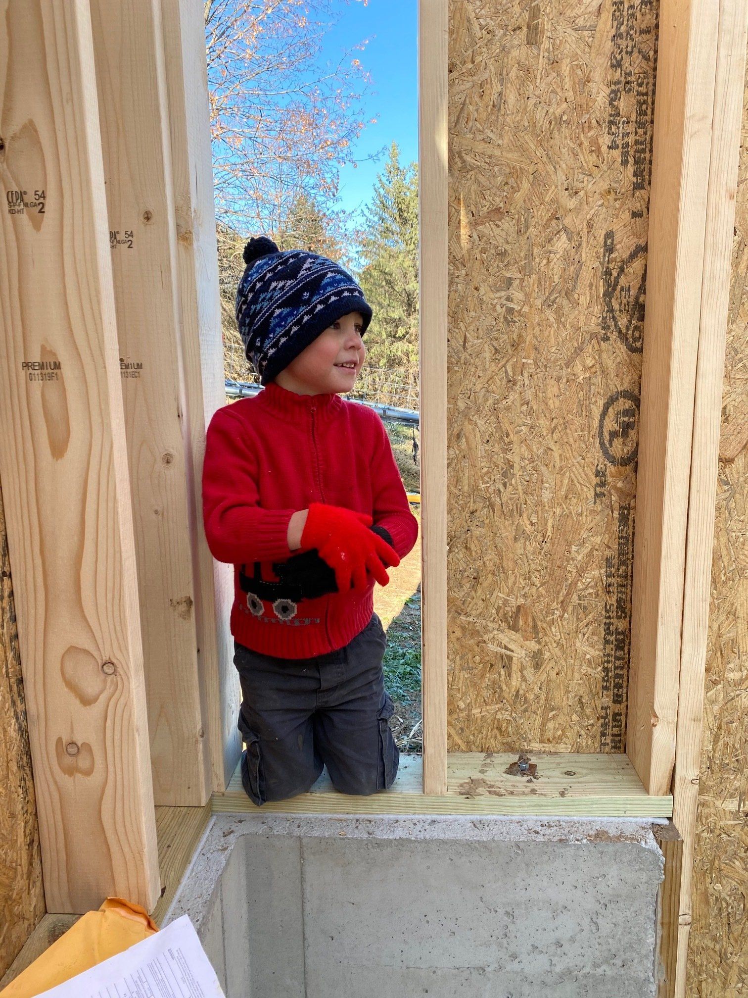 Young child in a red sweater and hat peers out a window frame during construction.