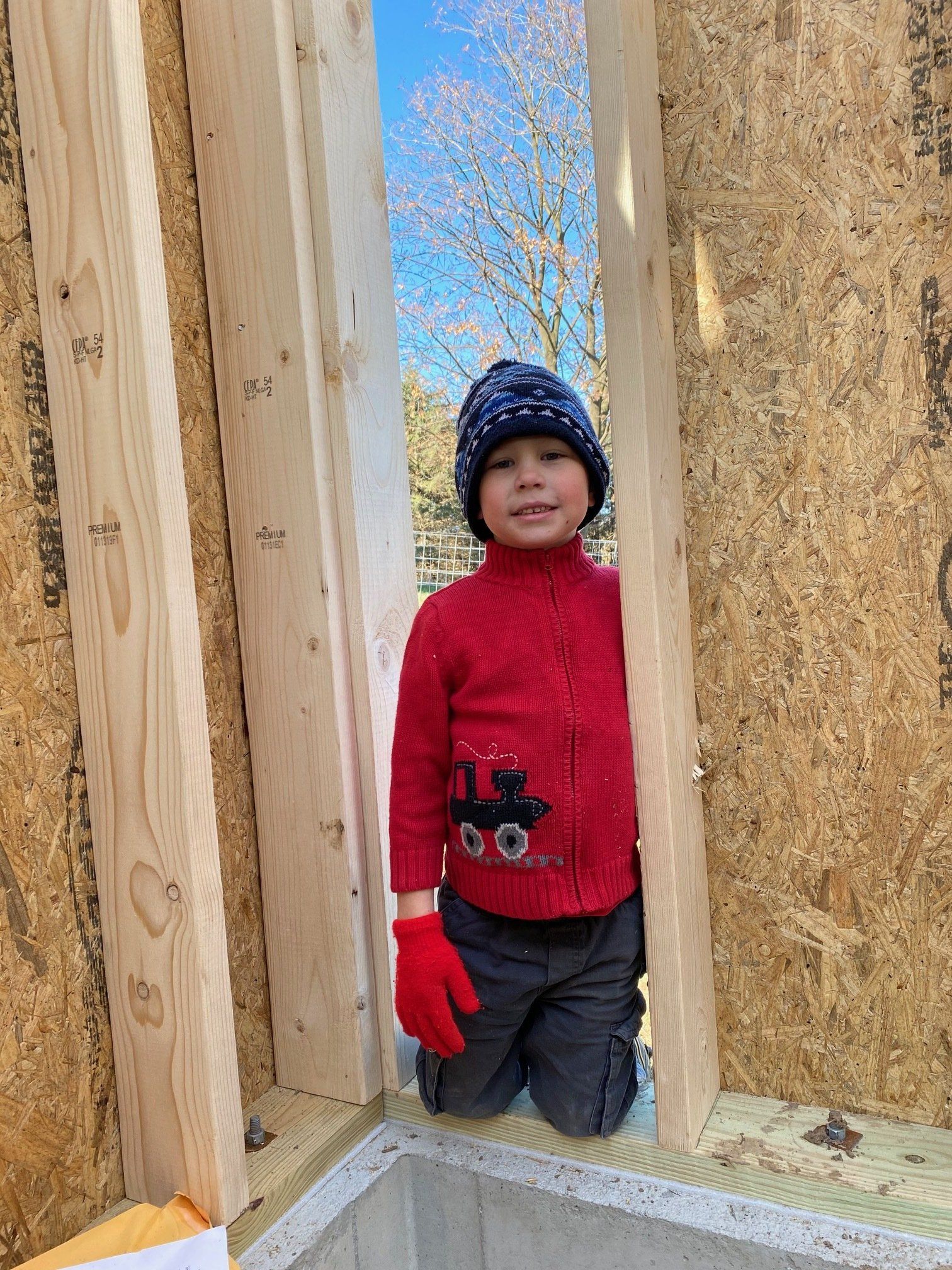 Child in red sweater, blue hat, and red gloves, peeks through a wooden wall opening; outdoor setting.