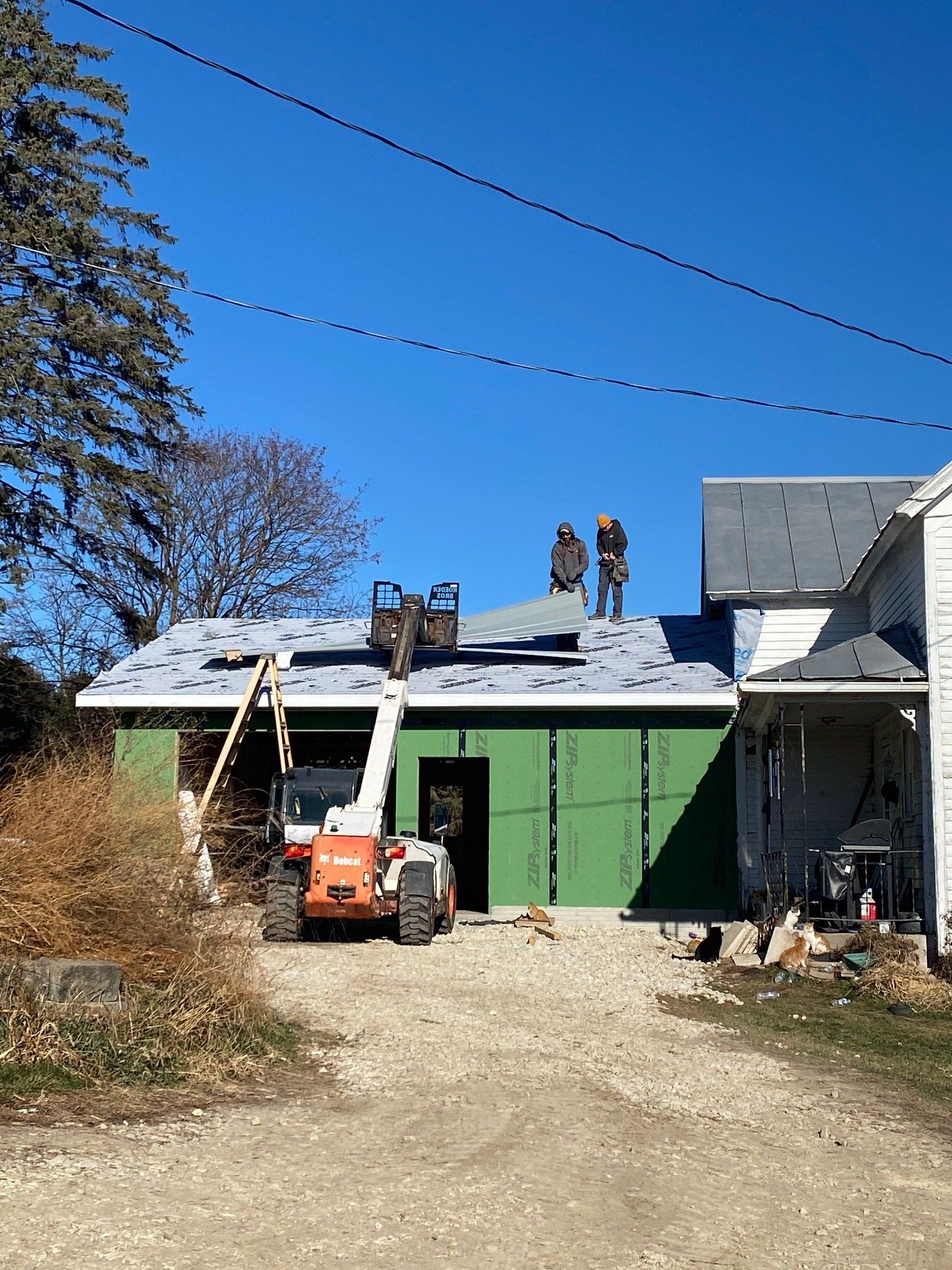 Construction workers on a roof installing sheet metal, using a lift. Green siding, blue sky, and a dirt driveway.