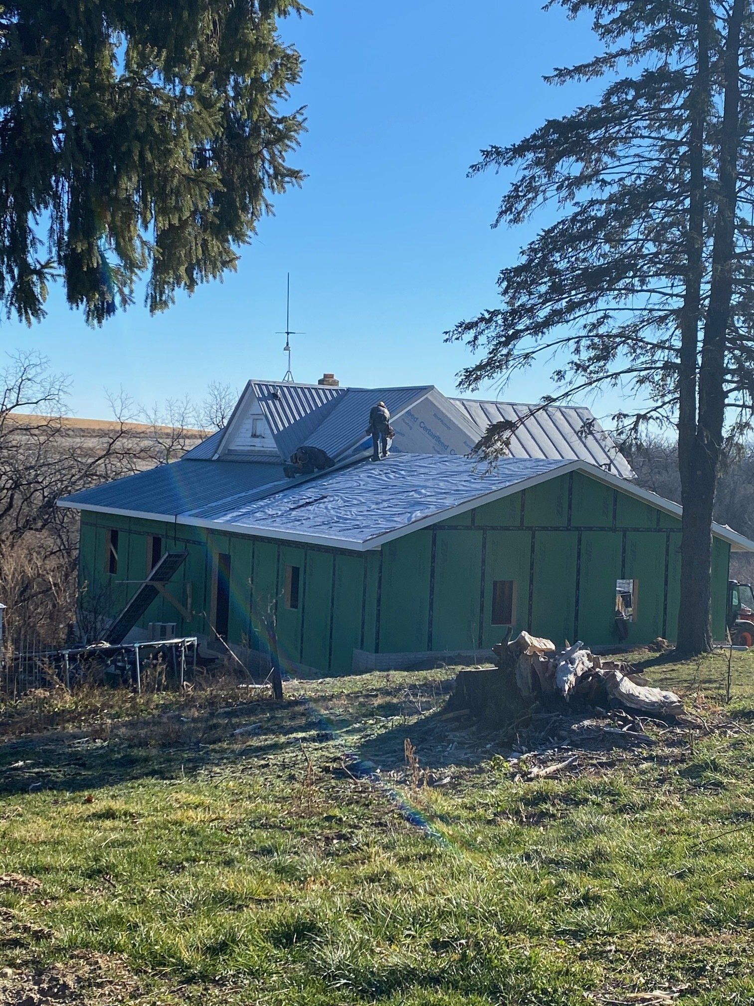 House under construction with green siding, workers on roof, trees, and blue sky.
