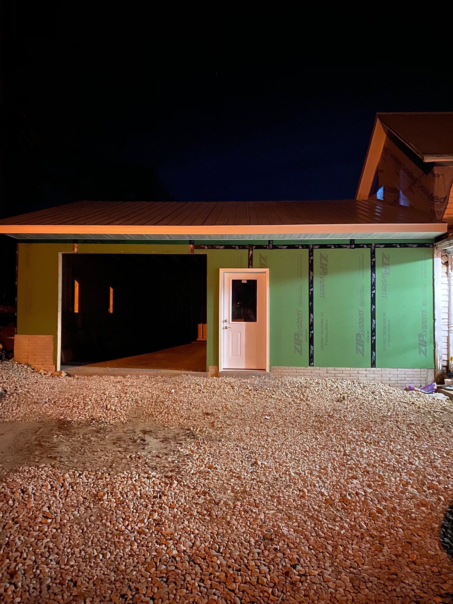 Unfinished garage exterior at night with open garage door, white door, gravel. Green siding, dark sky.