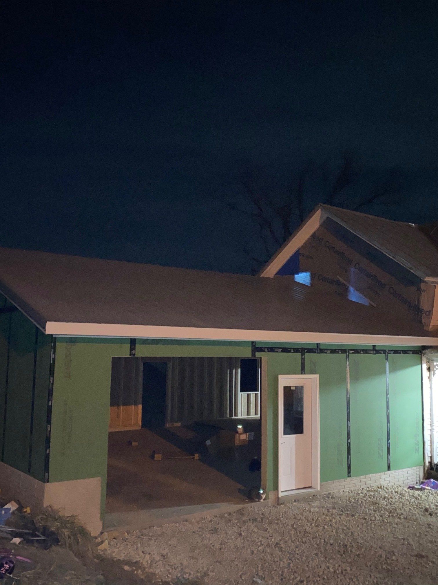 Construction of a building exterior at night. Green siding, white door, and gravel ground.