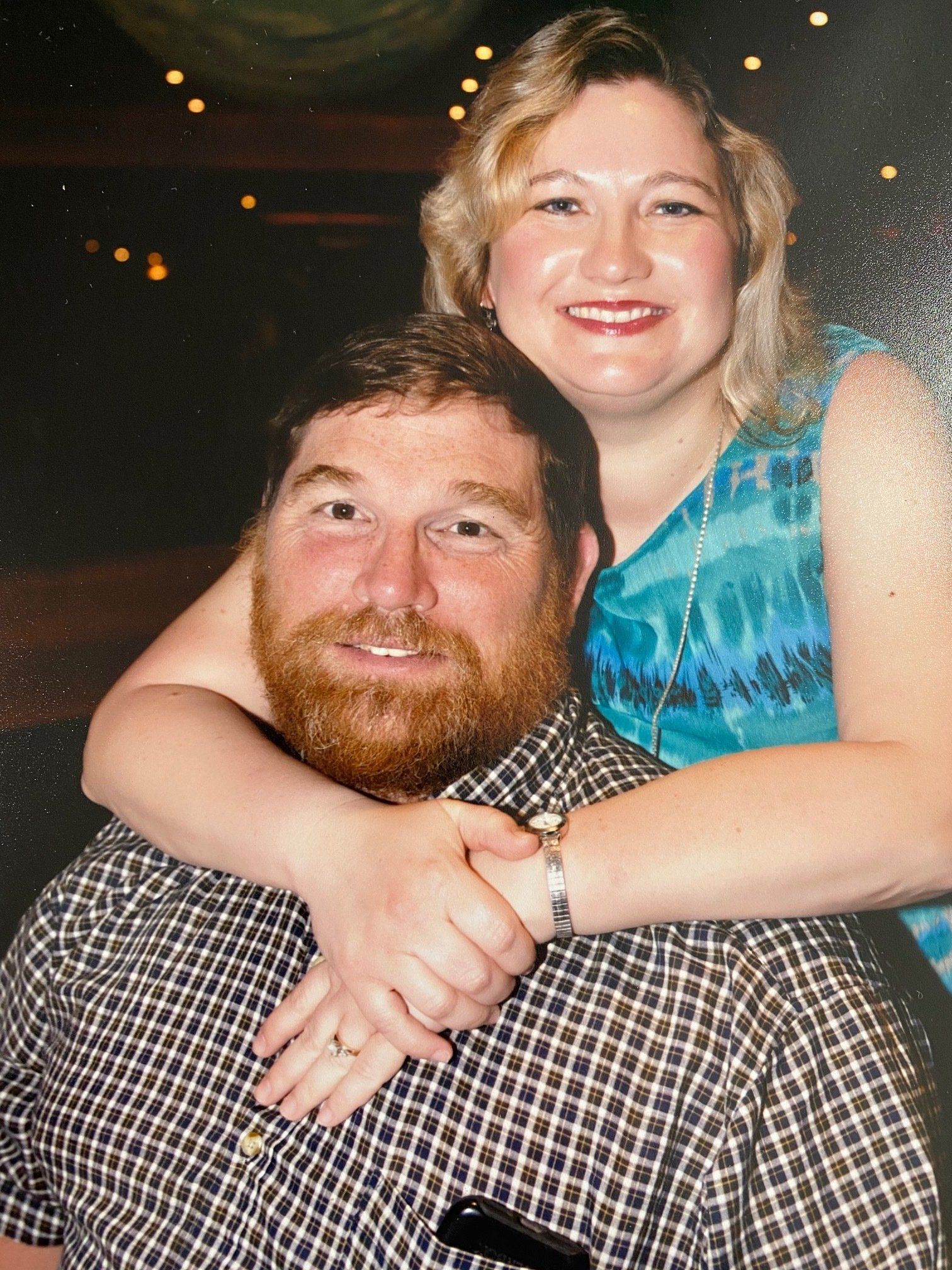 Couple embracing, man with beard in patterned shirt, woman in blue tie-dye top, smiling.