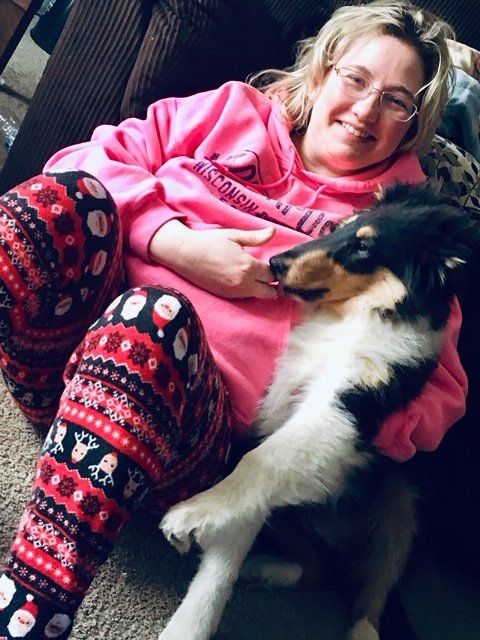 Woman in pink hoodie and festive pants cuddling a tri-color dog on a couch.