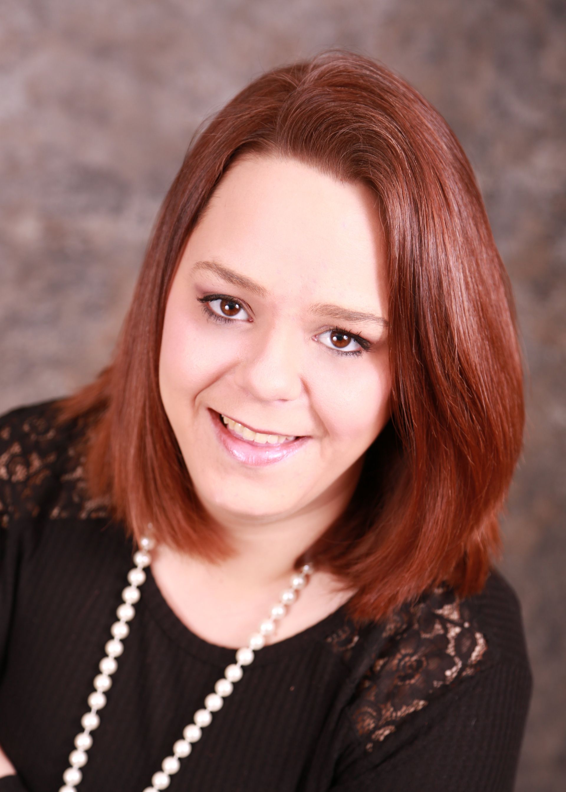 Smiling woman with shoulder-length auburn hair, wearing a black top and pearl necklace against a textured gray background