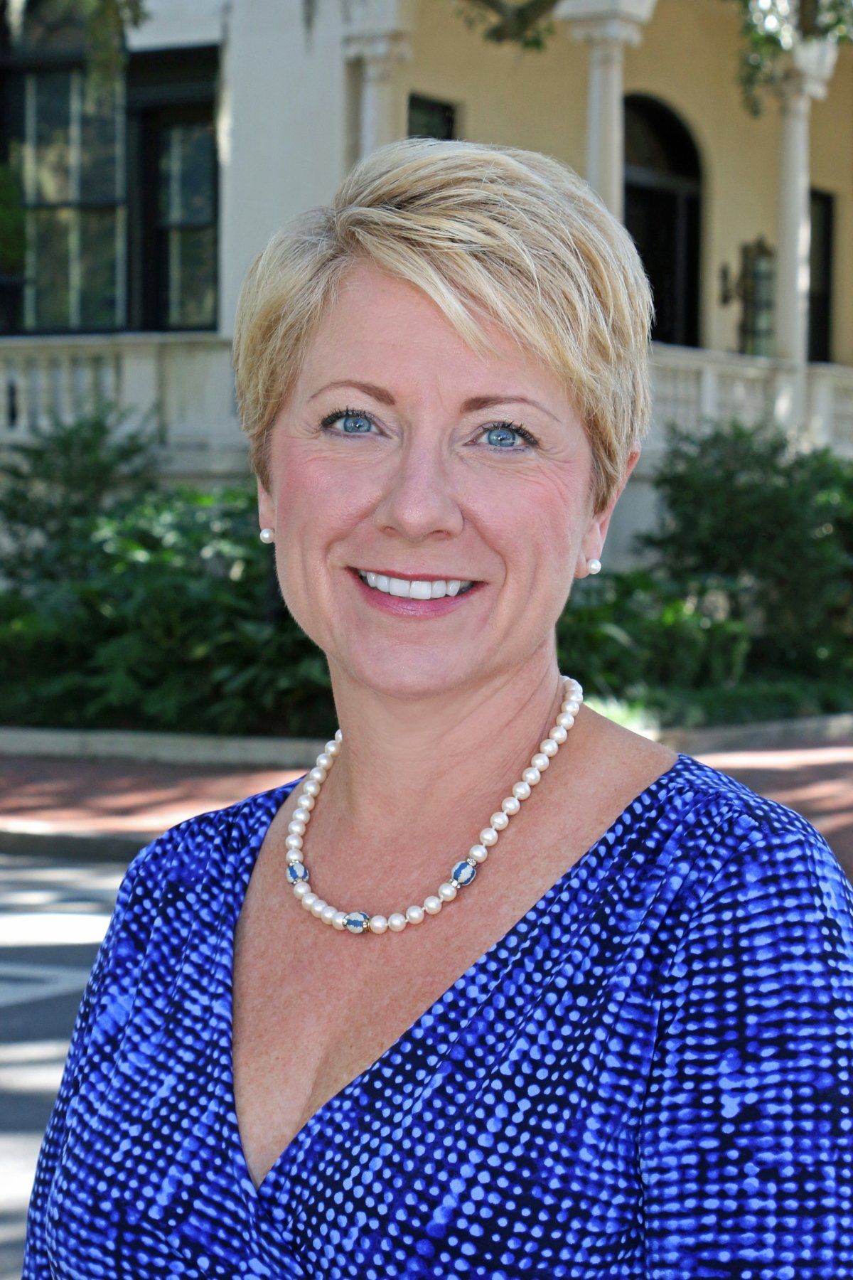 Smiling woman in a blue patterned dress and pearl necklace standing outdoors in front of a building