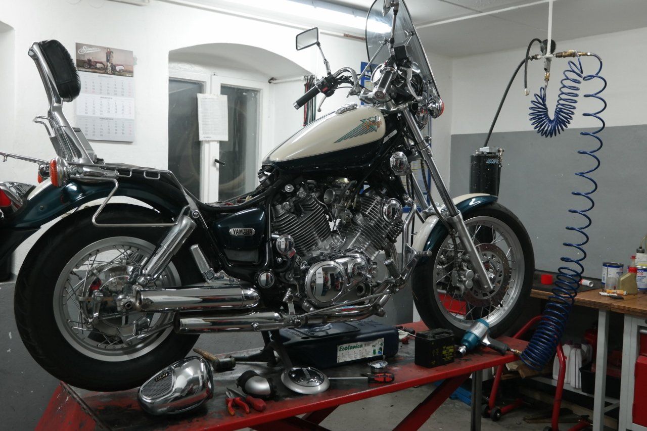 In einer Werkstatt steht ein Motorrad auf einer roten Hebebühne; in der Nähe befinden sich Ersatzteile und ein Luftschlauch.