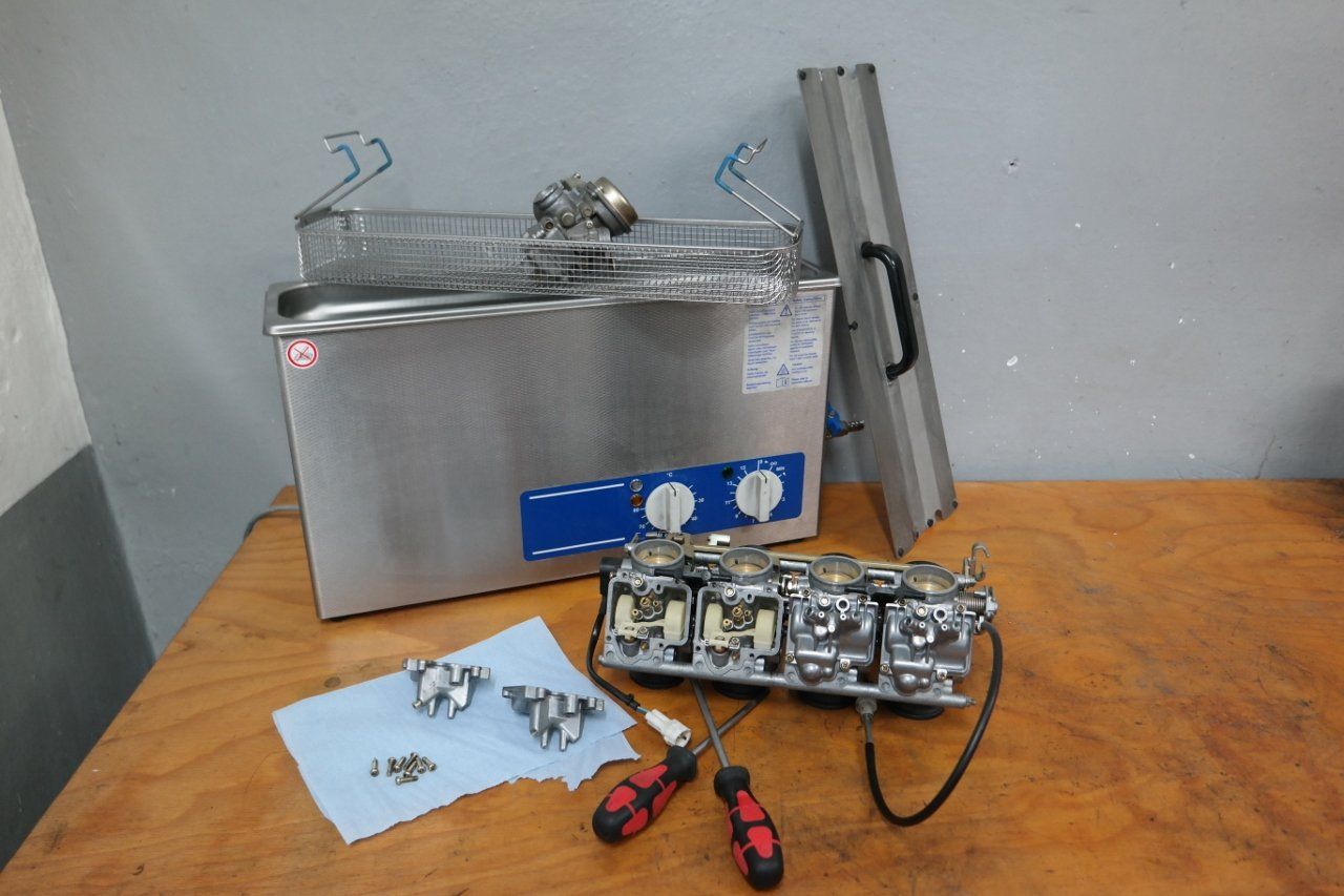 Ein Ultraschallreiniger, ein zerlegter Vergaserhalter für Motorräder und Schraubendreher liegen auf einer hölzernen Werkbank.