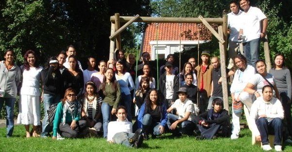 Een groep mensen staat voor een schommel in een grasveld met een gebouw met een rood dak.