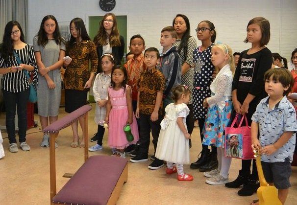 Een groep kinderen en volwassenen staat binnen, sommigen in traditionele kleding, met een bank ervoor.