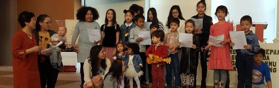 Een groep kinderen met papieren in hun handen zingt op het podium. Twee volwassenen staan ​​er vlakbij.