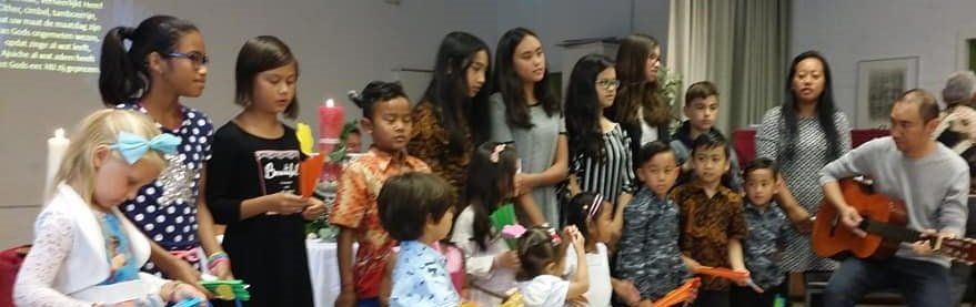Een groep kinderen en volwassenen zingt, begeleid door een man die gitaar speelt. Binnen, waarschijnlijk in een kerk.