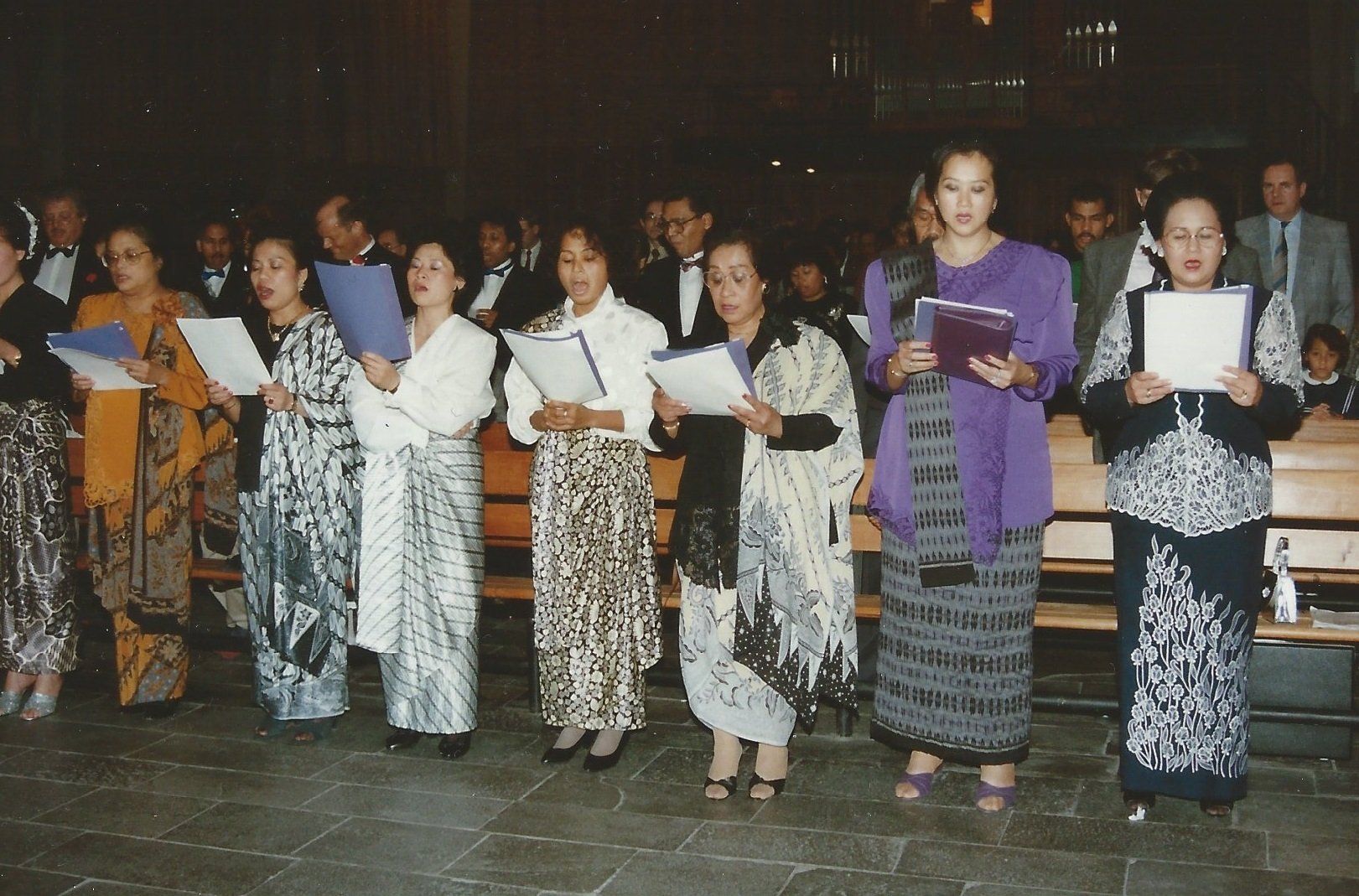 Een koor van mensen met bladmuziek zingt binnen. Ze dragen rokken met verschillende patronen, witte blouses en sjaals.