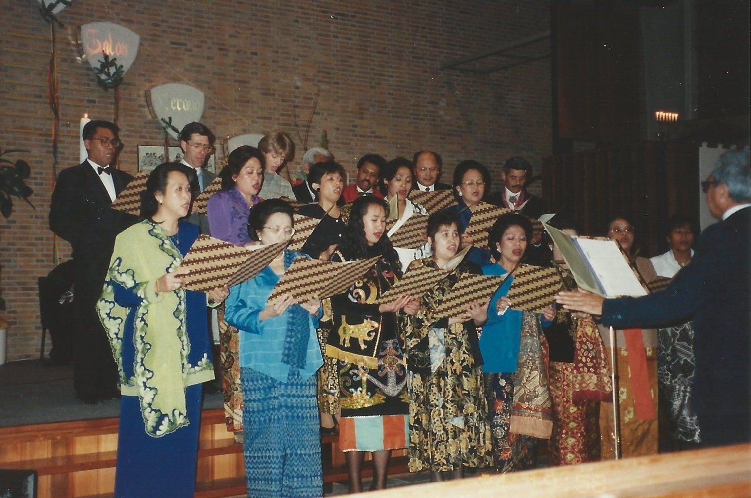 Een koor in traditionele kleding zingt, met liedbundels in de hand, onder leiding van een dirigent op het podium. Een bakstenen muur vormt de achtergrond.