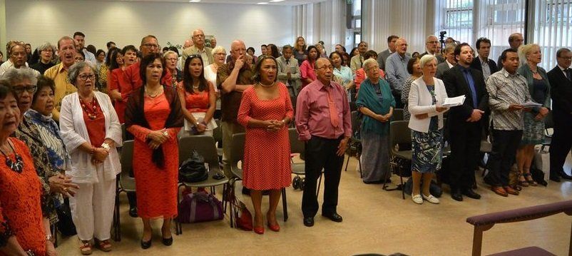 Mensen staan ​​binnen, sommigen in het rood, met de handen ineengeklemd, te luisteren.