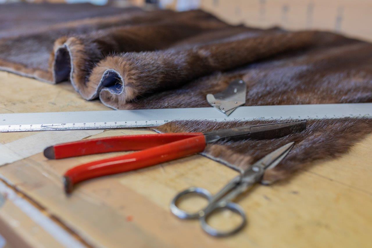 Su un banco da lavoro giace un pezzo di pelliccia marrone, accanto al quale si trovano un righello, delle pinze con il manico rosso e delle forbici argentate.