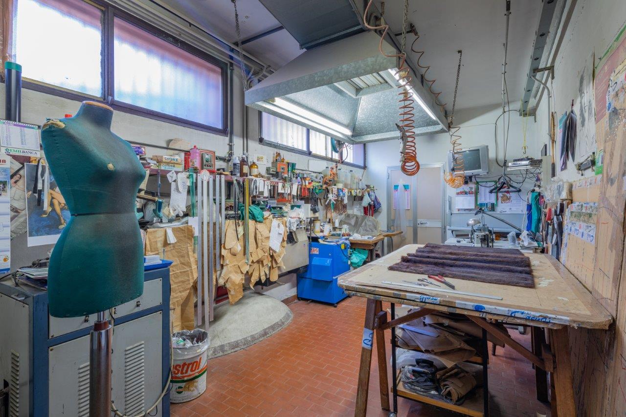 Un'officina disordinata con un manichino su un piedistallo, un grande banco da lavoro in legno, attrezzi e materiali appesi alle pareti.