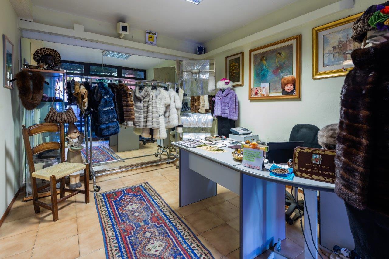 Interno di una pellicceria con bancone, sedia di legno e scaffali pieni di cappotti e cappelli di pelliccia in esposizione.
