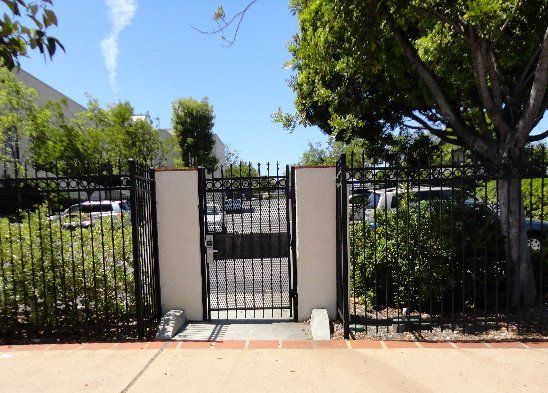 Black metal gate in a fence with concrete pillars. Trees and vehicles visible in background.