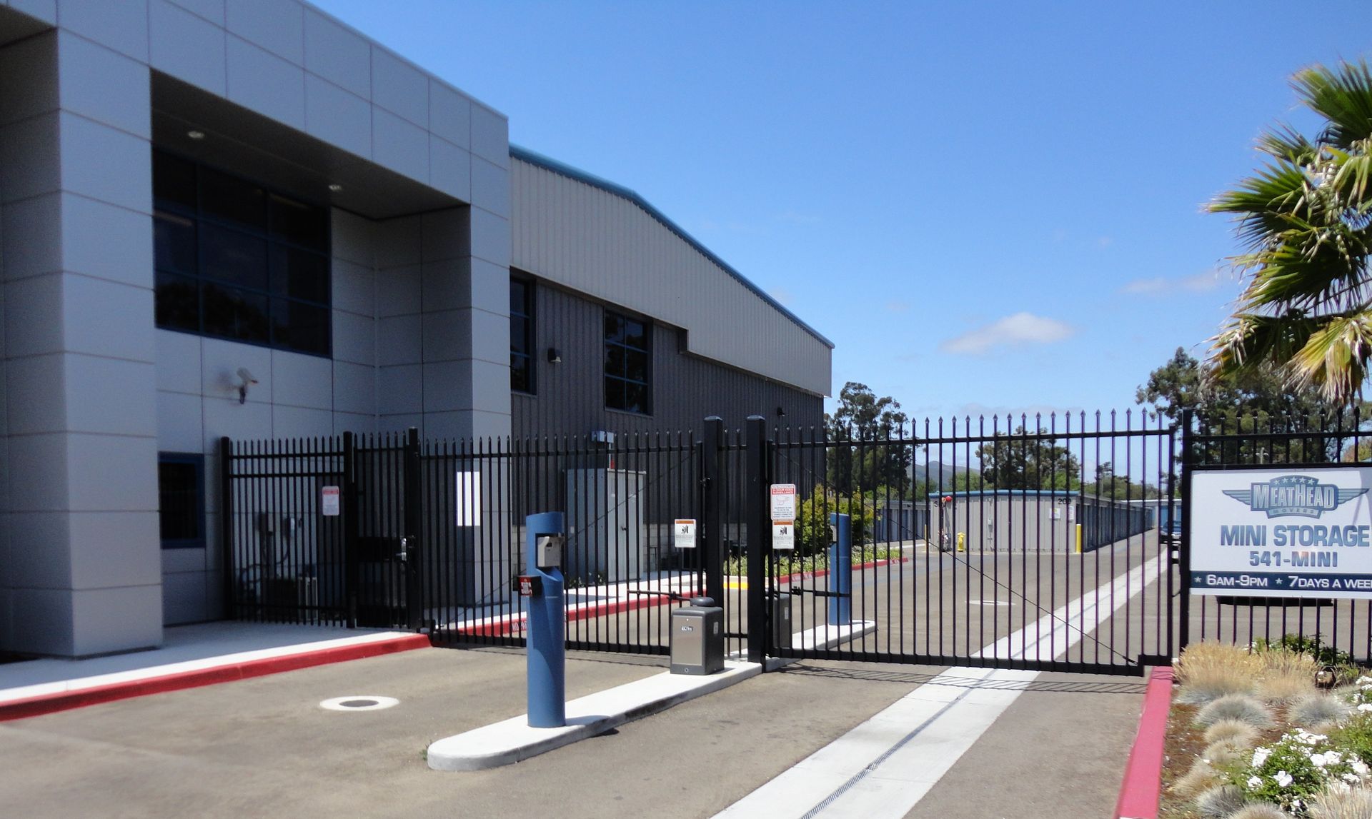 Secure storage facility with gated entrance, light-colored building, blue sky, and palm trees.