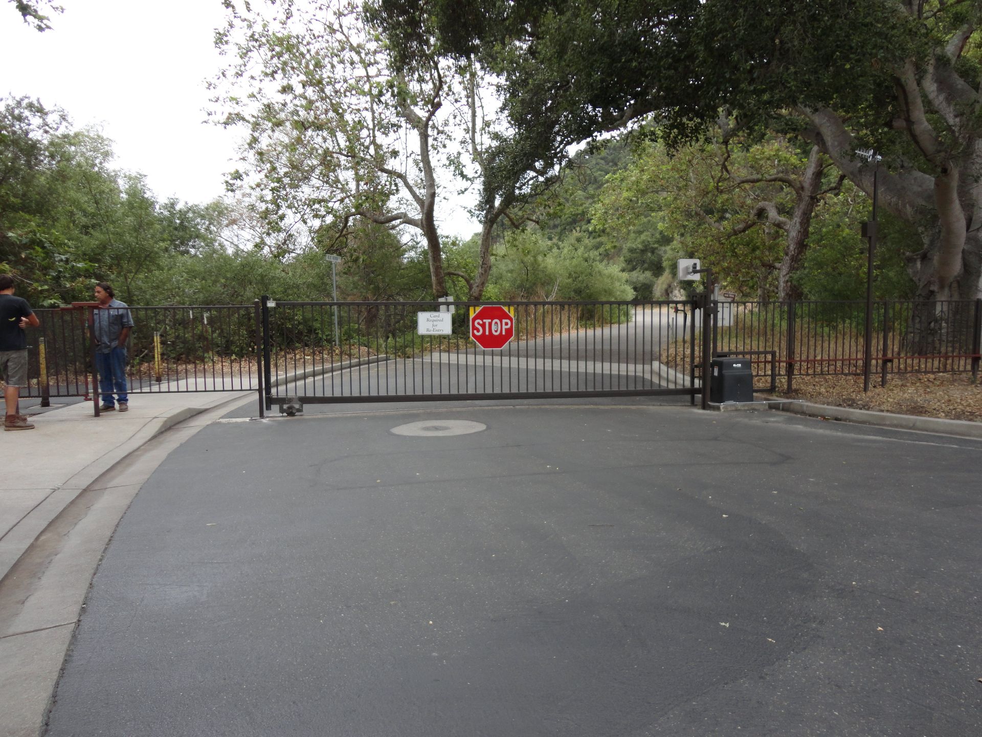 Black gate blocks a paved road, with a stop sign. Trees and people are on either side.