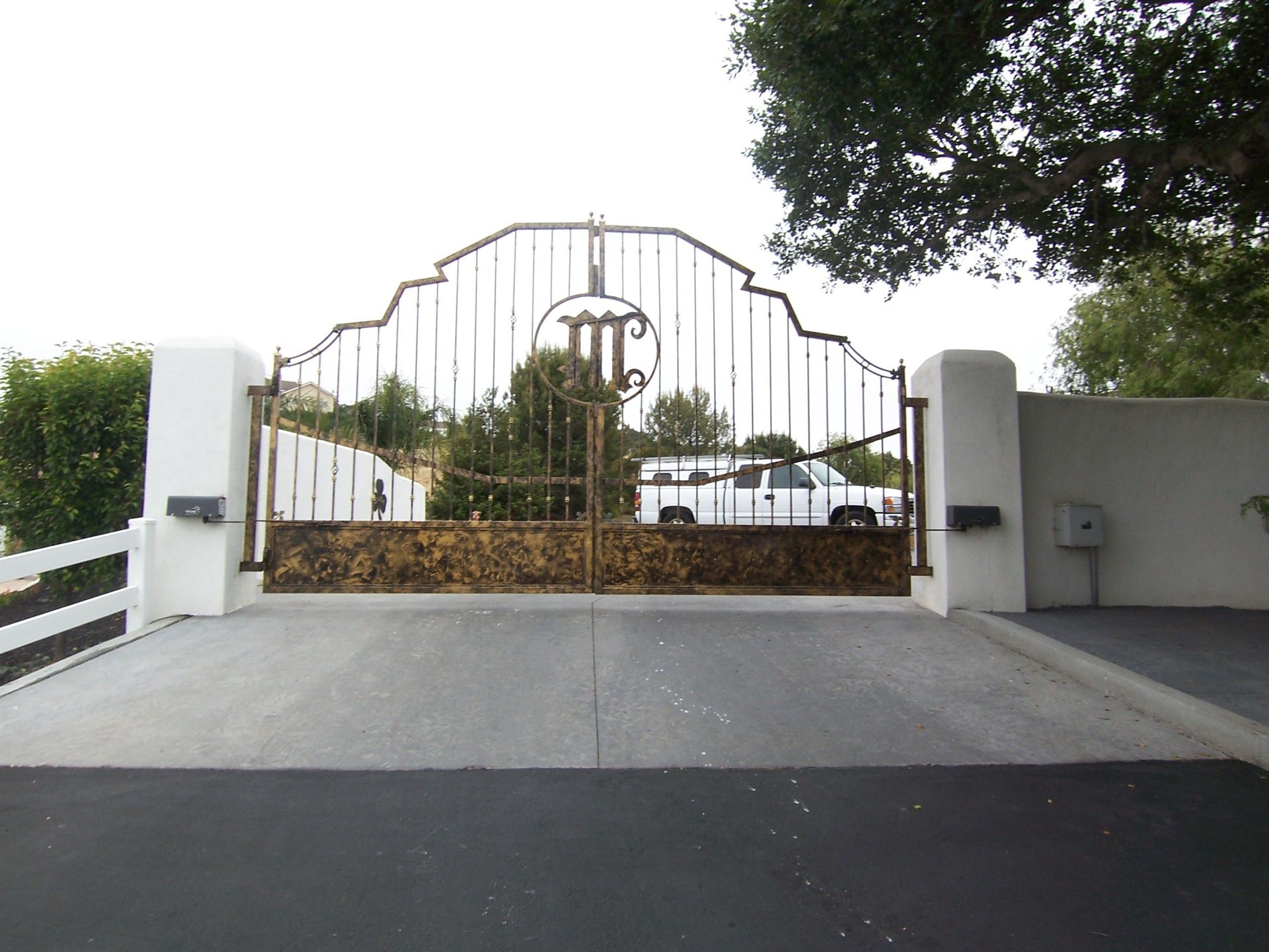 Ornate wrought iron gate with stone base, flanked by white pillars and walls. A white truck is visible behind the gate.