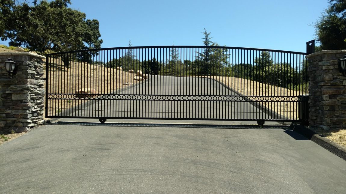 Black metal gate on asphalt driveway, flanked by stone pillars, against a sunny, tree-lined background.