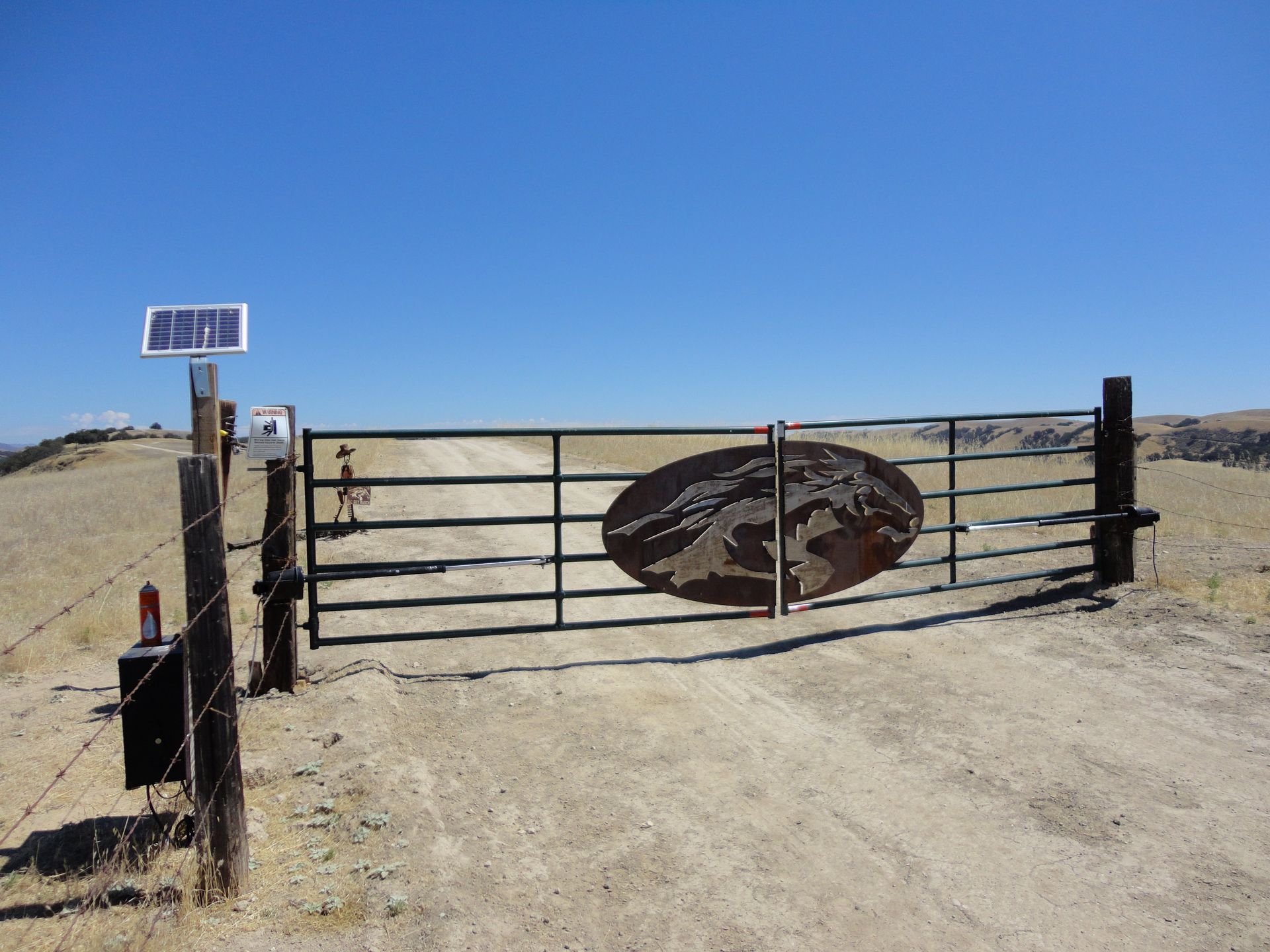 Metal gate with solar panel and mountain emblem, on dirt road with blue sky.