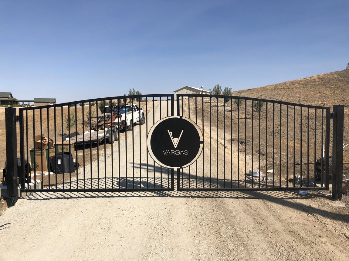 Black metal gate with logo, leading to a gravel driveway, with vehicles and a house visible.