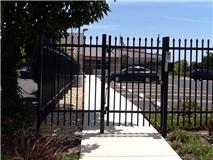 Black metal gate in front of a sidewalk leading to a building with a parking lot on a sunny day.