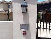 A beige stucco wall with a lantern, fire dept. box, and gate. A light-colored house is in the background.